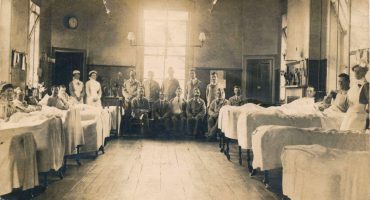 Patients in beds either side of ward and seated at the end with nurses