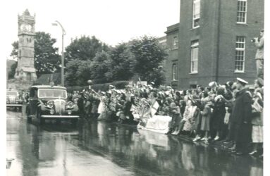 Crowds line street outside Infirmary waving flags, royal car passing by