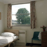 hospital bed in room, green plastic chair, wooden bedside cabinet, floral curtains, dark wood door with window pane