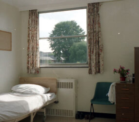 hospital bed in room, green plastic chair, wooden bedside cabinet, floral curtains, dark wood door with window pane
