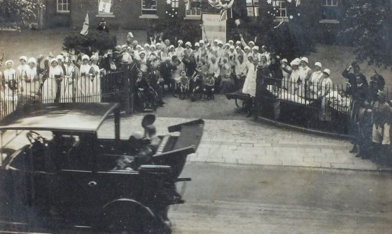 Old motor car driving past hospital staff and patients standing outside Infirmary gates waving