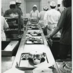 catering staff standing alongside conveyer belt loading trays with food