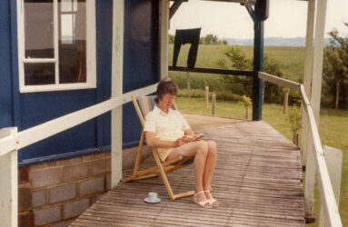 a colour photograph of person sat in deck chair on wooden walkway next to blue hut with white windows