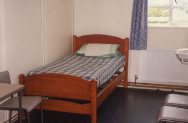a colour photo of bedroom with single pine bed and window with blue curtains