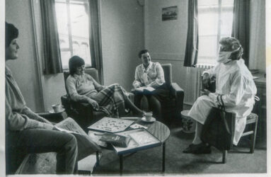 black and white photo of people seated in lounge with research visitor in white gown and face mask