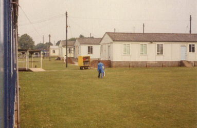 a colour photo of grass area and low white huts in distance