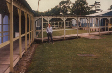 a colour photograph of grass area, wooden covered walkway between huts