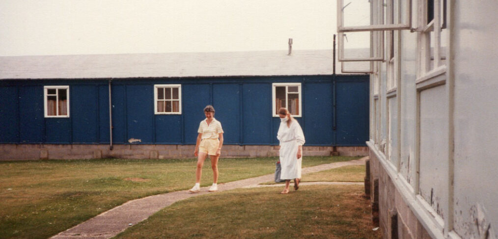 1 person in shorts and scientist in white gown and mask walk on grass next to hut