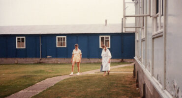 1 person in shorts and scientist in white gown and mask walk on grass next to hut