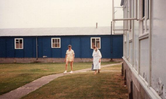 1 person in shorts and scientist in white gown and mask walk on grass next to hut