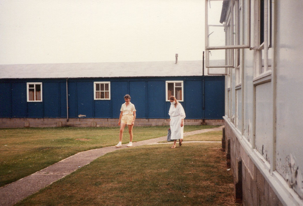 1 person in shorts and scientist in white gown and mask walk on grass next to hut