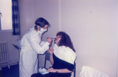 Colour photograph showing scientist in gown and mask giving nose drops to seated person