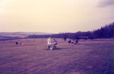 A colour photograph of grass area, a gowned figure stands in the middle with table
