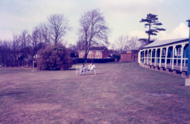 a colour photograph of grass area, 2 pople on bench and house in distance