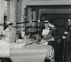 Princess with nurses talking to boy in traction on a bed in ward