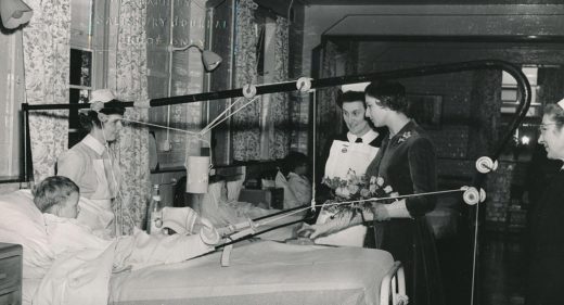 Princess with nurses talking to boy in traction on a bed in ward