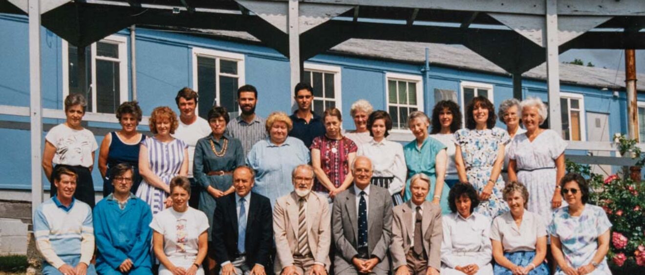colour photograph of a group of staff in 2 rows in front of blue building