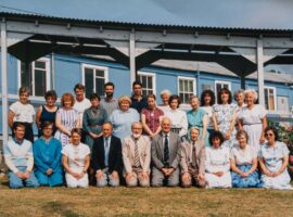 colour photograph of a group of staff in 2 rows in front of blue building
