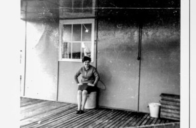 a black and white photo of woman seated on wooden walkway beside hut