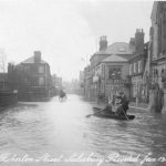 Three men in a rowing boat in flood water, horse and cart in the distance