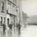 Soldier, two men and a boy in flat caps stand in flood water, larger crowd of people in distance by the clock tower