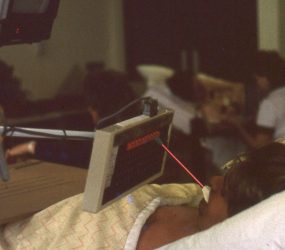 patient in bed using pencil with rubber end in mouth to tap onto a keyboard, monitor suspended overhead