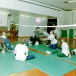 physiotherapists and occupational therapists sit on floor with patients some in wheelchairs, some on the floor playing volleyball in gym