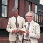 two men standing behind microphone, in front of the Spinal Unit building