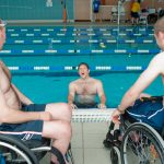 two men in wheelchairs at poolside looking at a third man in the pool hanging on to the side